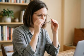 Femme inspectant de près des lunettes achetées en ligne, assise à un bureau en bois dans un bureau à domicile